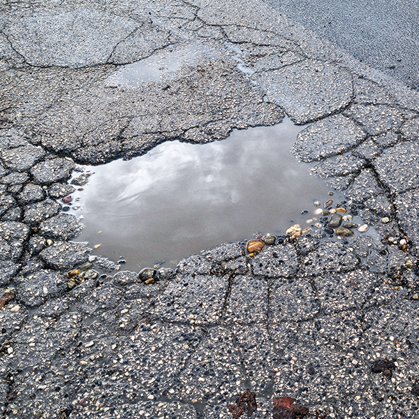A high-angle close-up captures a large, water-filled pothole reflecting a cloudy sky, surrounded by a network of deep alligator cracks and loose gravel on a deteriorating asphalt surface. This gritty texture image highlights issues of infrastructure neglect and roadway erosion, making it highly relevant for municipal repair reports, construction news, and urban decay backgrounds.